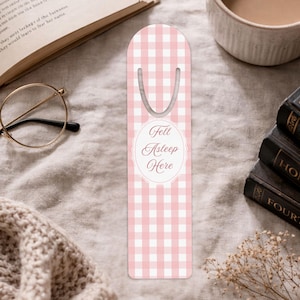 May include: A pink and white checked bookmark with a silver loop at the top. The bookmark features the text "Fell Asleep Here" in a decorative oval. The bookmark is placed near books, a cup of tea, and spectacles.