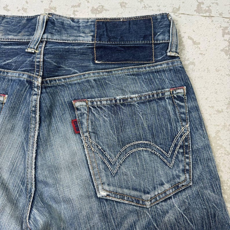 May include: Close-up of the back of blue denim jeans. The jeans have a faded wash with white stitching details on the back pocket. A small red tag is visible on the pocket, and a dark blue leather patch is at the waistband.