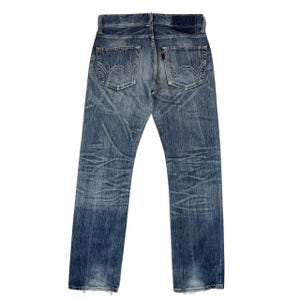 May include: Faded blue denim jeans with a classic five-pocket design. The jeans show signs of wear and tear, with distressed areas and a red tag on the back pocket. The back view highlights the stitching and the overall vintage aesthetic.