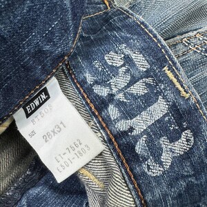 May include: Close-up of a pair of blue denim jeans. The jeans feature a white tag with the brand "EDWIN" and size "28x31". The jeans have a faded design on the back pocket area.