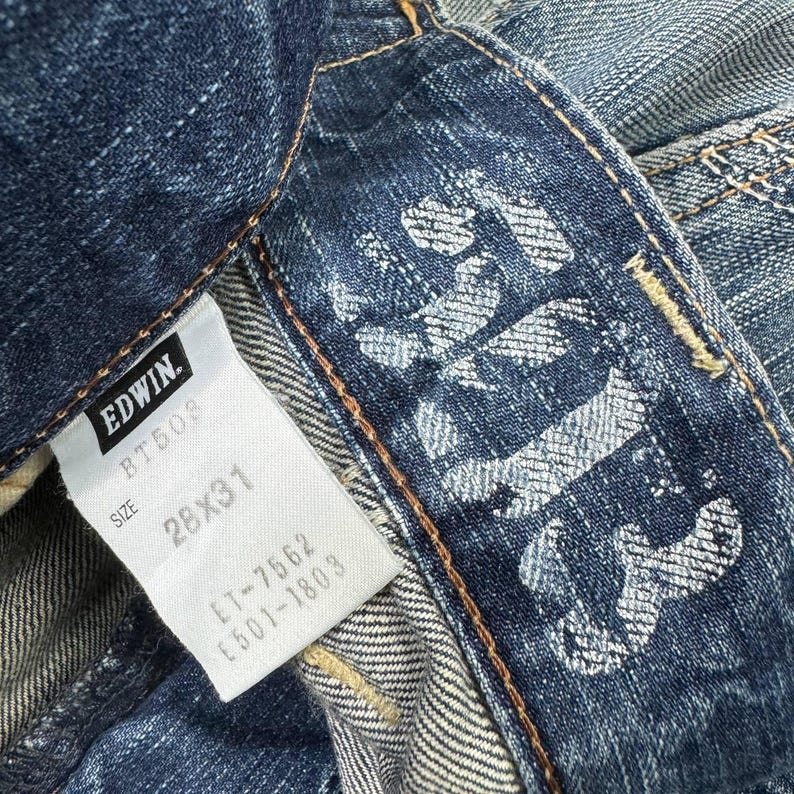 May include: Close-up of a pair of blue denim jeans. The jeans feature a white tag with the brand "EDWIN" and size "28x31". The jeans have a faded design on the back pocket area.