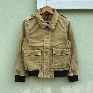 Puede incluir: Chaqueta de pana color canela con cuello, dos bolsillos delanteros y cierre de botones. La chaqueta tiene un dobladillo y puños negros acanalados. La chaqueta está colgada en una percha de madera sobre un fondo verde claro.