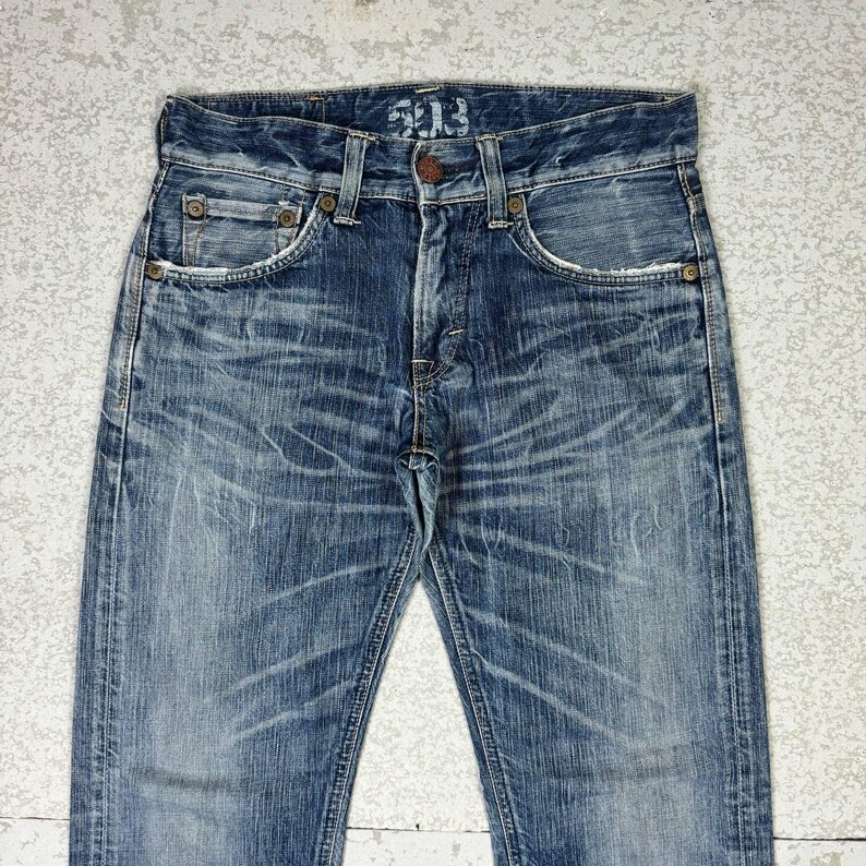 May include: Faded blue denim jeans with a distressed look. The jeans have a classic five-pocket design, a button fly, and visible wear patterns. The top of the jeans has the number "203" printed on it.