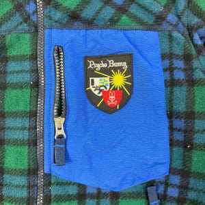 May include: A blue, green, and black plaid fleece jacket with a blue pocket and a black zipper. The pocket features a Psycho Bunny logo patch with a sun, bunny, and shield design.