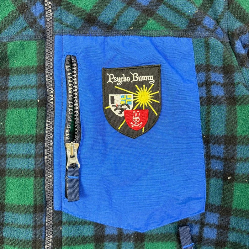 May include: A blue, green, and black plaid fleece jacket with a blue pocket and a black zipper. The pocket features a Psycho Bunny logo patch with a sun, bunny, and shield design.