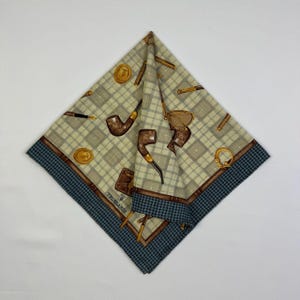 Puede incluir: Un pañuelo cuadrado plegado con un estampado de cuadros beige y gris, con pipas marrones, plumas y monedas doradas. El borde es un estampado de pata de gallo azul oscuro. La marca "TRUSSARDI" es visible en el pañuelo.