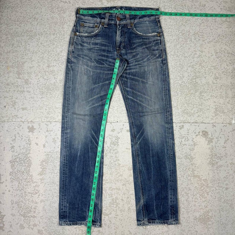 May include: Faded blue denim jeans with a button fly and classic five-pocket design. A green measuring tape shows the waist is approximately 15 inches across. The jeans have a worn, distressed look.