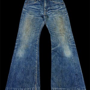 Puede incluir: Vaqueros vintage de mezclilla azul con diseño de pernera acampanada. Los vaqueros muestran signos de desgaste y decoloración, con un diseño clásico de cinco bolsillos y cierre de botón y cremallera. La mezclilla tiene un lavado medio con zonas más claras.