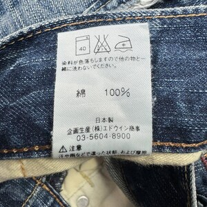 May include: Close-up of a denim jeans label with Japanese text and care symbols. The label indicates the jeans are 100% cotton and made in Japan. Washing instructions are also included.
