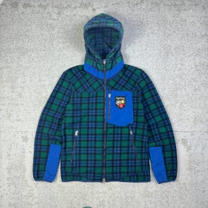 May include: A hooded fleece jacket in a green and blue plaid pattern. The jacket features a blue pocket with a patch, blue accents, and a full zipper. The hood and collar are also blue. The jacket has two side pockets with zippers.