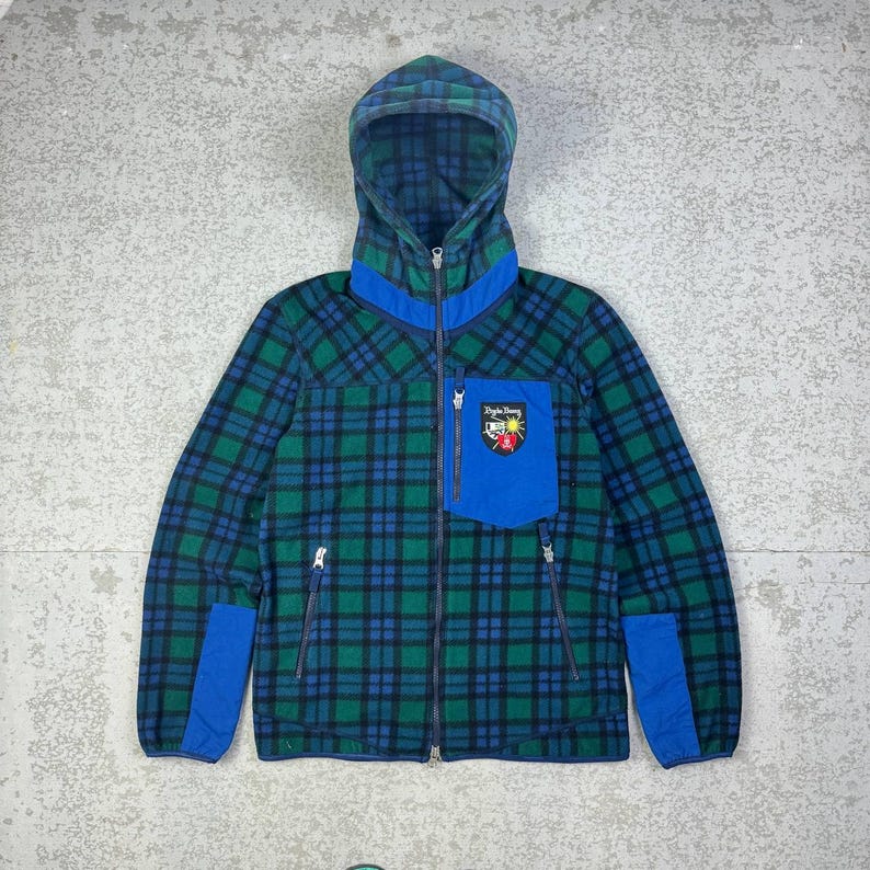 May include: A hooded fleece jacket in a green and blue plaid pattern. The jacket features a blue pocket with a patch, blue accents, and a full zipper. The hood and collar are also blue. The jacket has two side pockets with zippers.