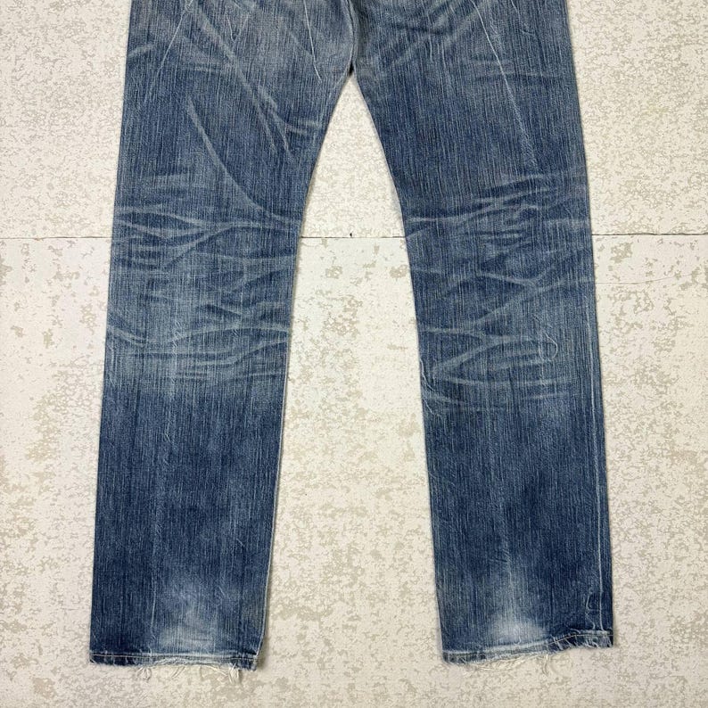 May include: Faded blue denim jeans with a straight leg cut. The jeans show signs of wear with visible fading and creasing throughout the fabric. The bottom hems are slightly frayed, adding to the vintage aesthetic.
