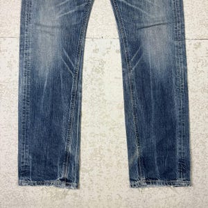 May include: Faded blue denim jeans with distressed details. The jeans have a straight leg cut and show signs of wear and tear, with visible fading and subtle whiskering. The hemline is slightly frayed.