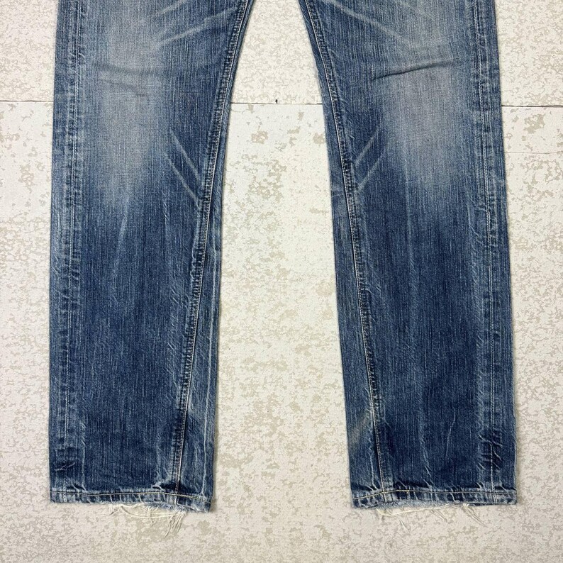 May include: Faded blue denim jeans with distressed details. The jeans have a straight leg cut and show signs of wear and tear, with visible fading and subtle whiskering. The hemline is slightly frayed.