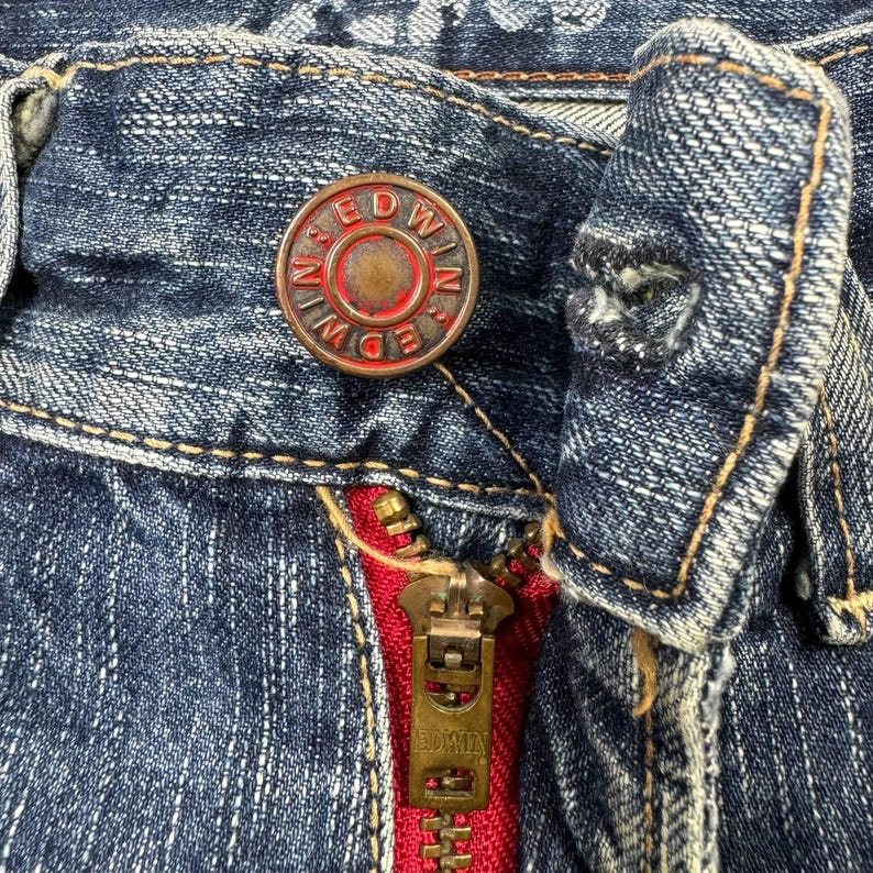 May include: Close-up of blue denim jeans with a red zipper and a metal button. The button is circular with the word "EDWIN" around the edge. The zipper pull is also metal and has the word "EDWIN" on it.