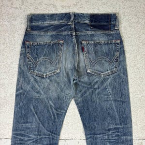 May include: Faded blue denim jeans with classic five-pocket styling. Features include a leather patch, red stitching accents, and the iconic back pocket design. The jeans have a worn, vintage look.