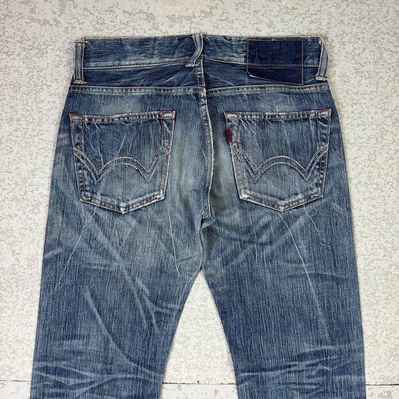 May include: Faded blue denim jeans with classic five-pocket styling. Features include a leather patch, red stitching accents, and the iconic back pocket design. The jeans have a worn, vintage look.
