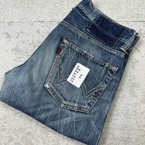 May include: Blue denim jeans with a faded wash and distressed details. Features a classic five-pocket design, a back pocket with a unique stitch pattern, and a red logo tag. The jeans have a folded appearance.