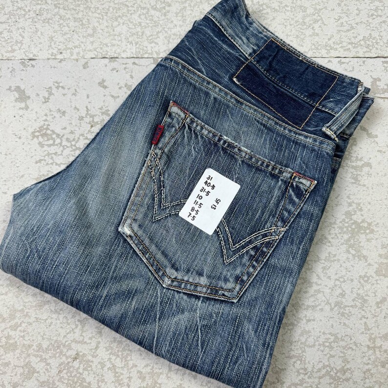 May include: Blue denim jeans with a faded wash and distressed details. Features a classic five-pocket design, a back pocket with a unique stitch pattern, and a red logo tag. The jeans have a folded appearance.