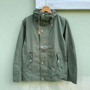 Puede incluir: Chaqueta con capucha verde oliva con cremallera y cierre de botones. La chaqueta tiene dos bolsillos delanteros y una capucha con cordón. Cuelga de una percha de madera contra una pared verde claro.