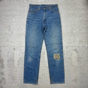 Könnte beinhalten: Hellblaue Jeans mit einem Katzenaufnäher am Knie. Die Jeans haben ein klassisches Fünf-Taschen-Design und einen Knopf- und Reißverschluss. Der Katzenaufnäher ist beige mit grünen Augen.