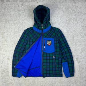 May include: A hooded jacket with a green and blue plaid pattern. The jacket has a blue interior, a blue pocket, and a zipper closure. A patch with text and a crest is on the pocket.