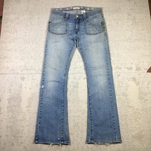 Puede incluir: Vaqueros de mezclilla azul claro con diseño de pernera acampanada. Los vaqueros tienen dos bolsillos delanteros y dos traseros. Hay salpicaduras de pintura blanca en la parte delantera de los vaqueros. Los vaqueros están planos sobre una superficie gris.