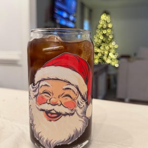 May include: A clear glass can-shaped cup filled with a dark drink and ice cubes. The cup has a smiling Santa Claus face design in red, white, and pink. A Christmas tree with blurred lights is in the background.