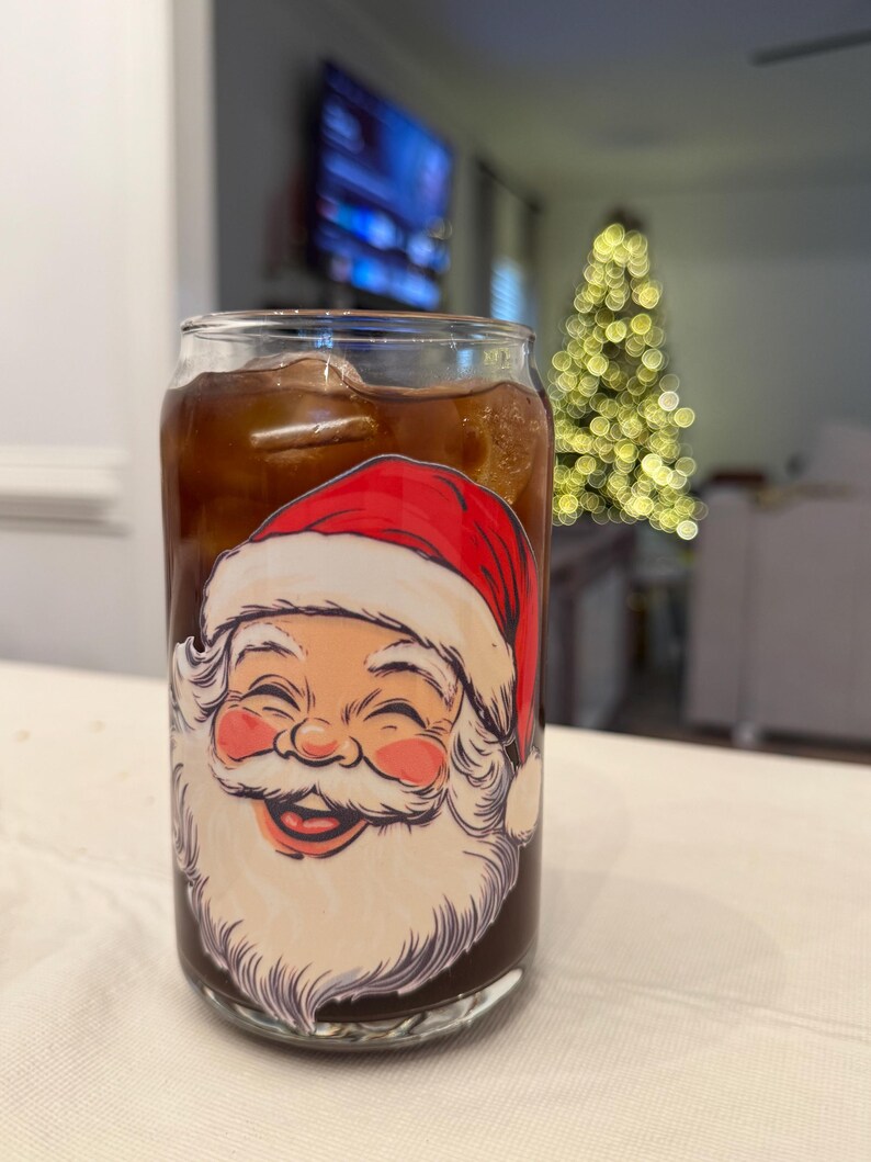 May include: A clear glass can-shaped cup filled with a dark drink and ice cubes. The cup has a smiling Santa Claus face design in red, white, and pink. A Christmas tree with blurred lights is in the background.