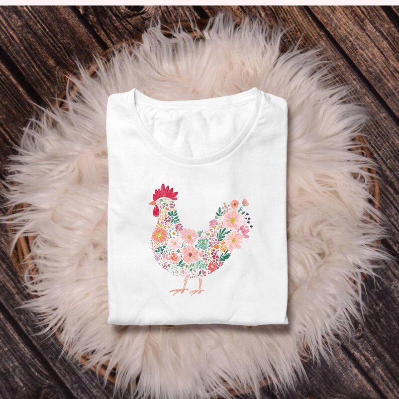 May include: White t-shirt featuring a floral hen design. The hen is composed of various colorful flowers, with a red comb and wattle. The shirt is folded and placed on a fluffy, white surface, with a wooden background.