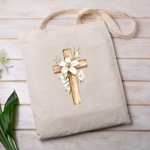 May include: Beige tote bag featuring a watercolor illustration of a wooden cross adorned with white lilies and greenery. The cross is centrally positioned, creating a religious or Easter theme. The bag has a simple design with a neutral color palette.