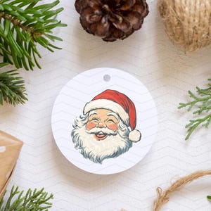 May include: A circular Christmas decoration with a smiling Santa Claus illustration. The design features a red hat, white beard, and rosy cheeks. The background includes festive elements like pine needles, a pine cone, and twine.