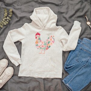 May include: A light gray hoodie with a floral rooster design. The rooster is composed of pink, yellow, and green flowers. The hoodie has a front pocket, a hood, and long sleeves. A pair of blue jeans and white sneakers are next to the hoodie.