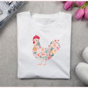 May include: White t-shirt featuring a floral hen design. The hen is composed of various flowers in shades of pink, orange, and green, with a red comb and wattle. The shirt is folded and placed on a gray surface.