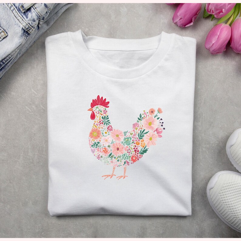 May include: White t-shirt featuring a floral hen design. The hen is composed of various flowers in shades of pink, orange, and green, with a red comb and wattle. The shirt is folded and placed on a gray surface.