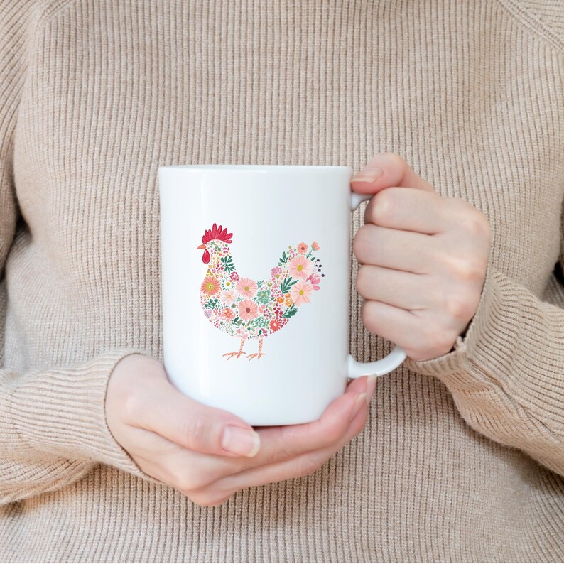 May include: White ceramic mug featuring a floral hen design. The hen is composed of various flowers in shades of pink, orange, and green. The mug has a large handle and is held by a person.