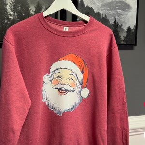 May include: Maroon sweatshirt with a smiling Santa Claus face, white beard, and red hat with a white pom-pom. The sweatshirt hangs on a white hanger. Framed landscape art is visible in the background.