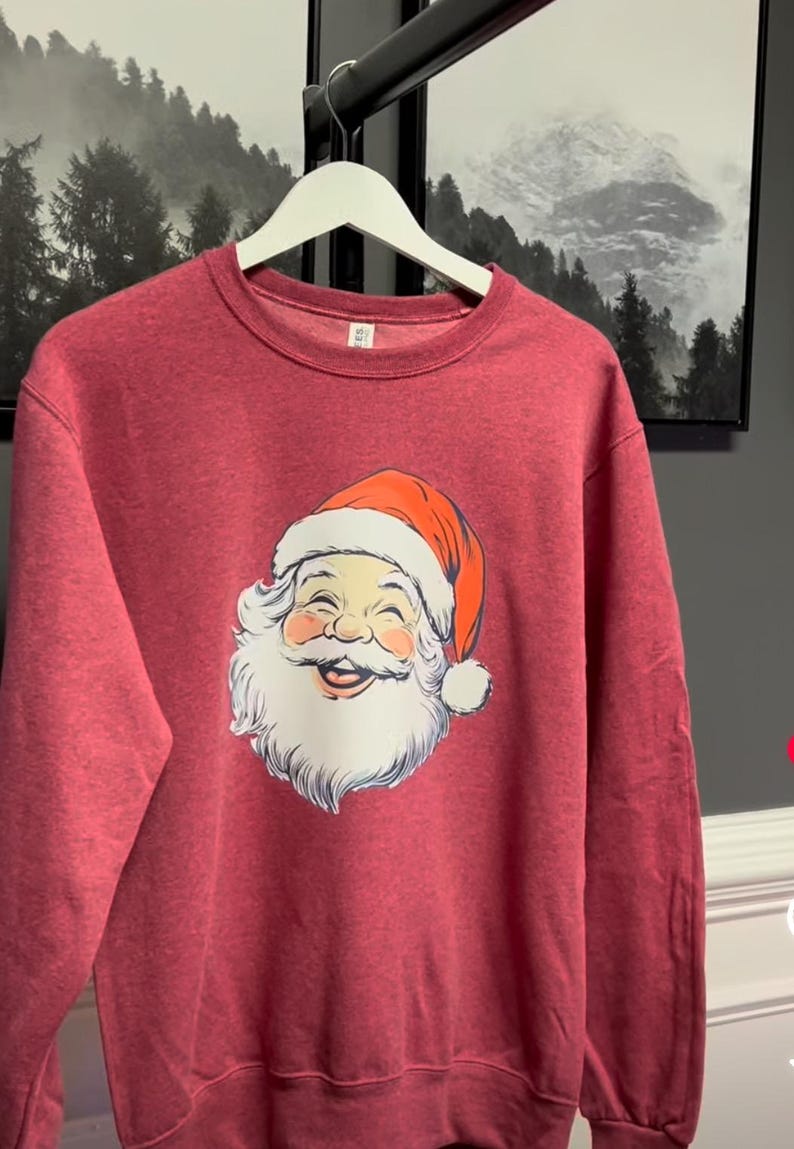 May include: Maroon sweatshirt with a smiling Santa Claus face, white beard, and red hat with a white pom-pom. The sweatshirt hangs on a white hanger. Framed landscape art is visible in the background.