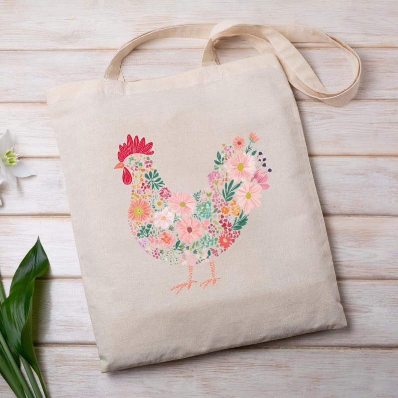May include: A beige canvas tote bag featuring a floral rooster design. The rooster is composed of various colorful flowers in shades of pink, orange, and green. The bag has a simple, long strap for carrying.
