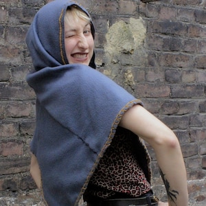 May include: A woman wearing a blue hooded cape with a brown trim. The cape is pulled up over her head and she is looking over her shoulder with a smile. She is wearing a leopard print shirt and black jeans.