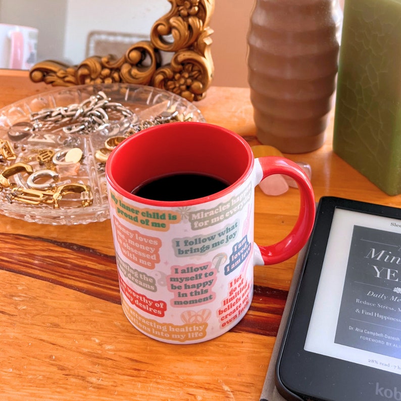 May include: A coffee mug with a red handle and interior, filled with dark liquid, displaying positive affirmations on colorful labels. It rests on a wooden surface beside an e-reader and a jewellery tray. The mug's interior is red.