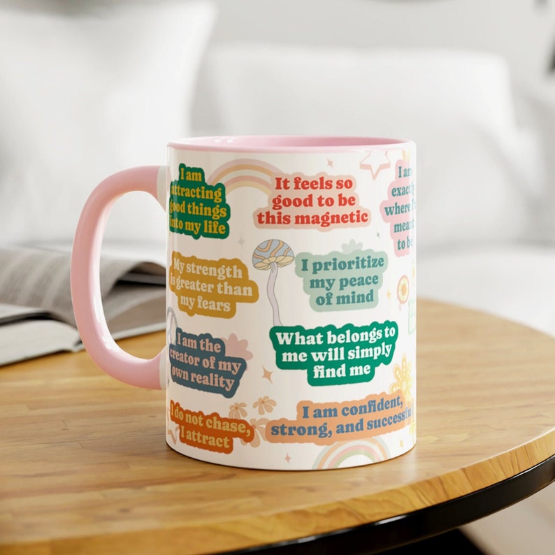 May include: A white ceramic mug with a pink handle and rim. The mug is decorated with colourful text stickers, including phrases like "I am attracting good things" and "I prioritise my peace of mind." The mug sits on a wooden table.