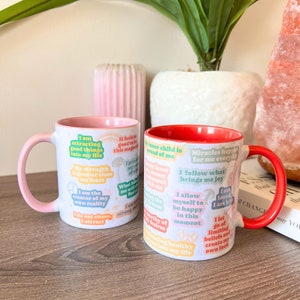 May include: Two white ceramic mugs with positive affirmations printed on them. One has a pink interior and handle, the other a red interior and handle. The mugs are decorated with colourful text stickers with inspirational quotes.