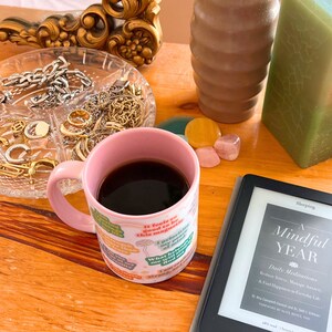 May include: A pink mug filled with dark coffee sits on a wooden surface next to a digital reader and a book titled "A Mindful Year". The mug has a colorful design with text. Jewellery and candles are in the background.
