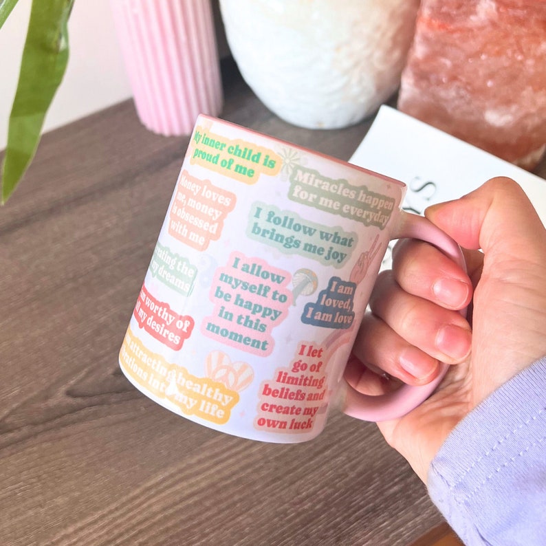 May include: A white ceramic mug with a pink handle and interior, adorned with colourful sticker-style affirmations. The affirmations include phrases such as "I allow myself to be happy in this moment" and "Miracles happen for me everyday."