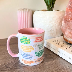 May include: A white ceramic mug with a pink interior and handle, featuring inspirational quotes in various colours. The mug has a gradient pink design and is placed on a wooden surface. The text includes phrases like "I am attracting good things into my life."