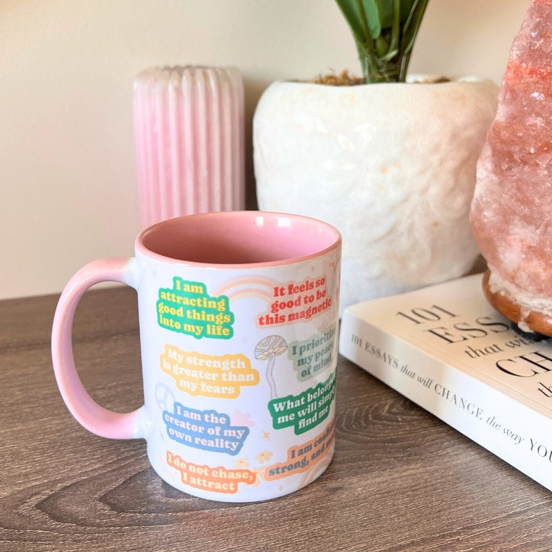 May include: A white ceramic mug with a pink interior and handle, featuring inspirational quotes in various colours. The mug has a gradient pink design and is placed on a wooden surface. The text includes phrases like "I am attracting good things into my life."