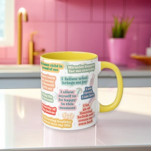 May include: A white ceramic mug with a yellow interior and handle. The mug features colourful text affirmations and positive statements. The mug is on a white surface with a pink background.