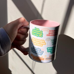 May include: White ceramic mug with a pink interior and handle. The mug is decorated with colourful text stickers that read, "I am attracting good things into my life," and other positive affirmations. The mug is held by a hand.