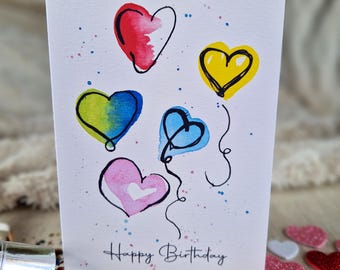 May include: A white card with a hand-drawn illustration of five colorful heart-shaped balloons. The balloons are in shades of red, yellow, green, blue, and pink. The card has the words "Happy Birthday" written in black ink at the bottom.